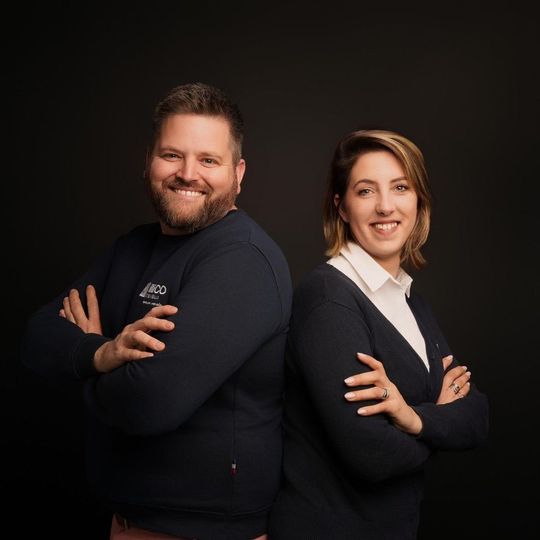 Un homme et une femme dos à dos, les bras croisés, sourient sur un fond noir.