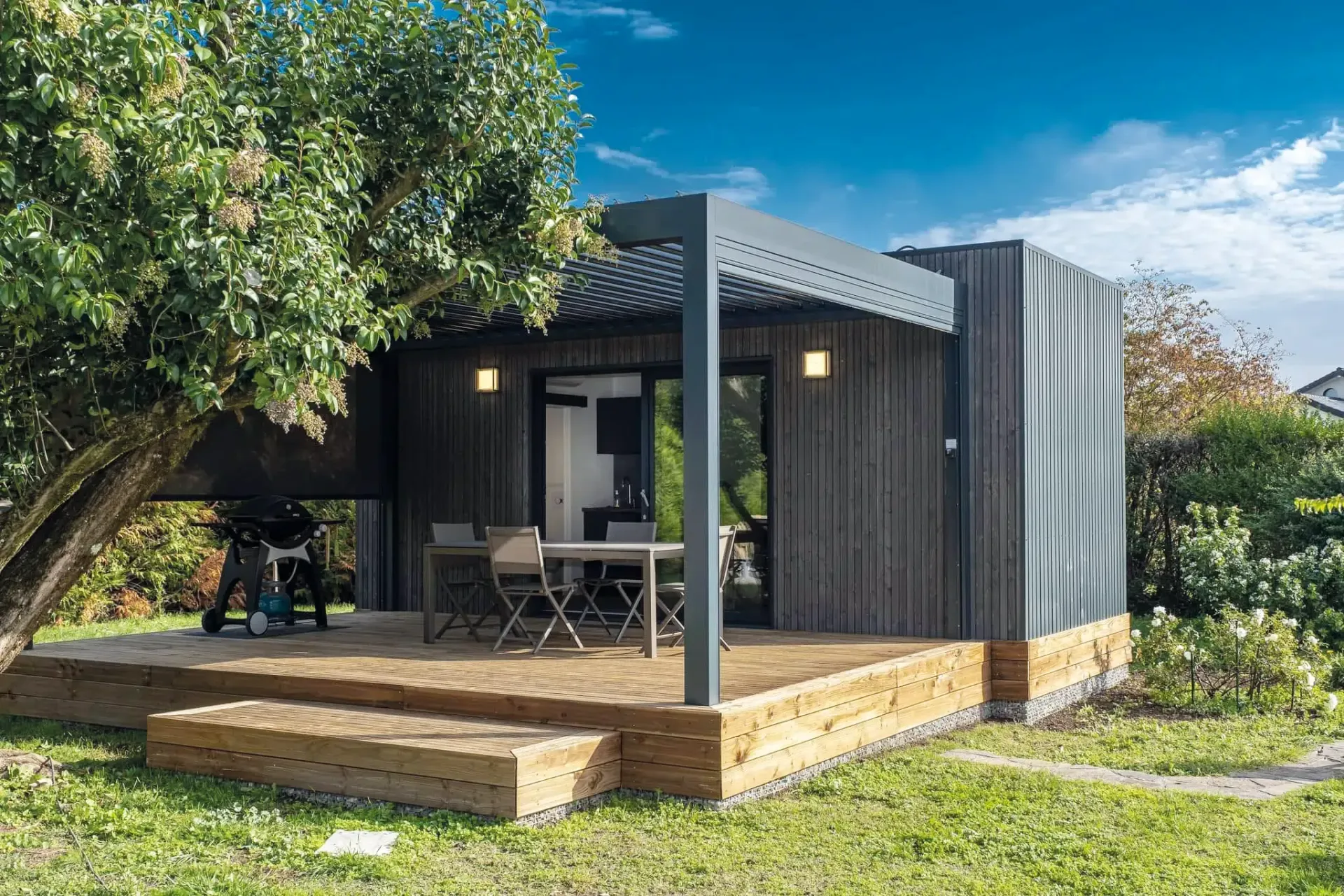 Petit chalet avec terrasse, coin salon extérieur et pergola. Bardage noir, terrasse en bois, pelouse verte.