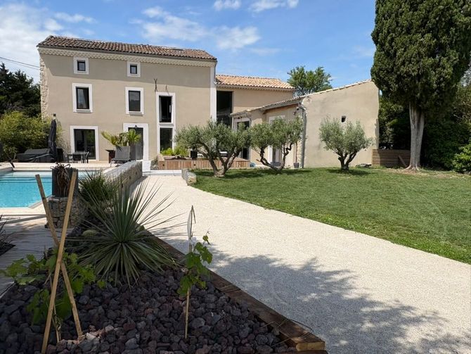 Vue extérieure d'une maison beige avec piscine, allée de gravier, oliviers et pelouse sous un ciel bleu.