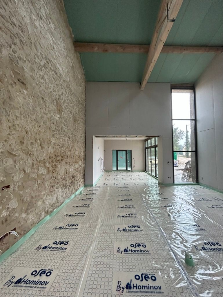 Intérieur d'un bâtiment en construction, montrant un mur en pierre, l'isolation du sol et des poutres en bois.