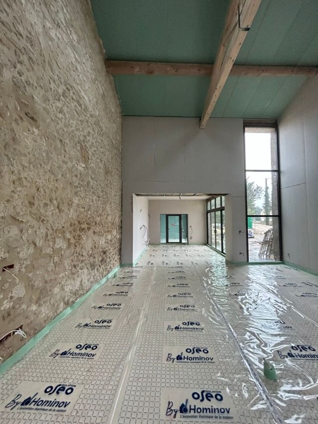 Intérieur d'un bâtiment en construction, montrant un mur en pierre, l'isolation du sol et des poutres en bois.