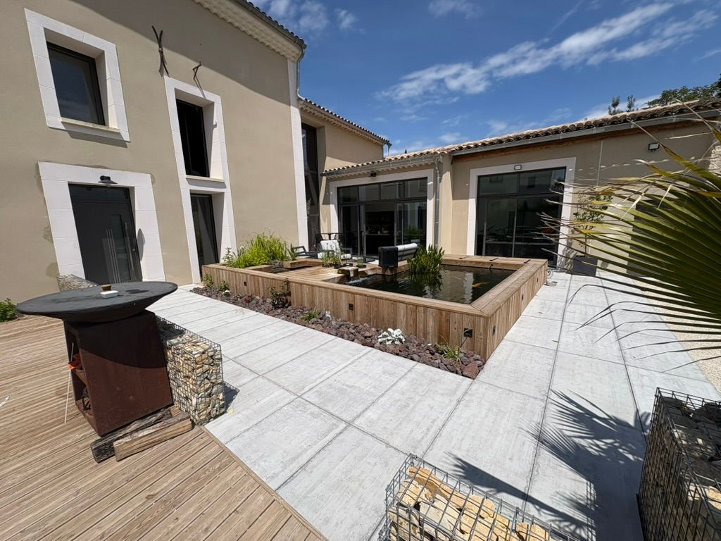Vue extérieure d'une maison beige avec une cour intérieure agrémentée d'un bassin surélevé en bois et d'une terrasse en pierre. Journée ensoleillée.