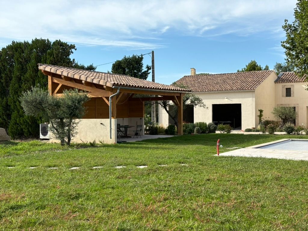Abri extérieur en bois avec toit de tuiles, soubassement en pierre et pelouse verte. Maison en arrière-plan.