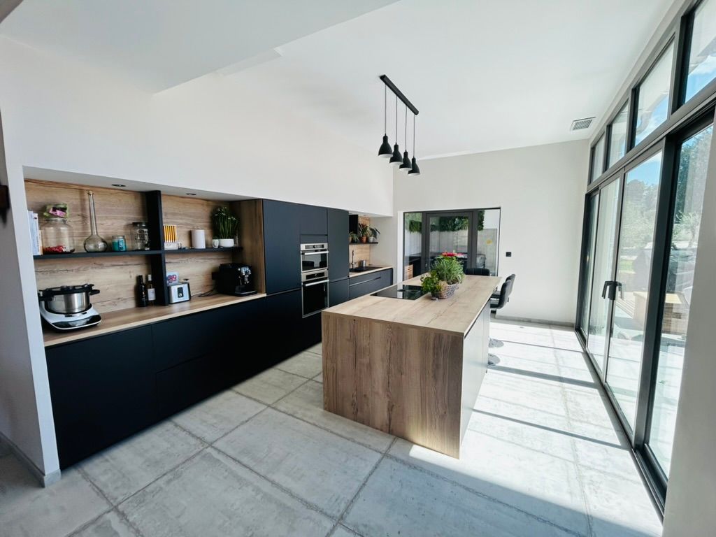 Cuisine moderne avec placards noirs et en bois, îlot central et grandes fenêtres.