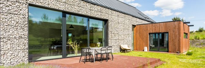 Maison moderne avec façade en pierre, grandes portes vitrées, terrasse extérieure et extension en bois.