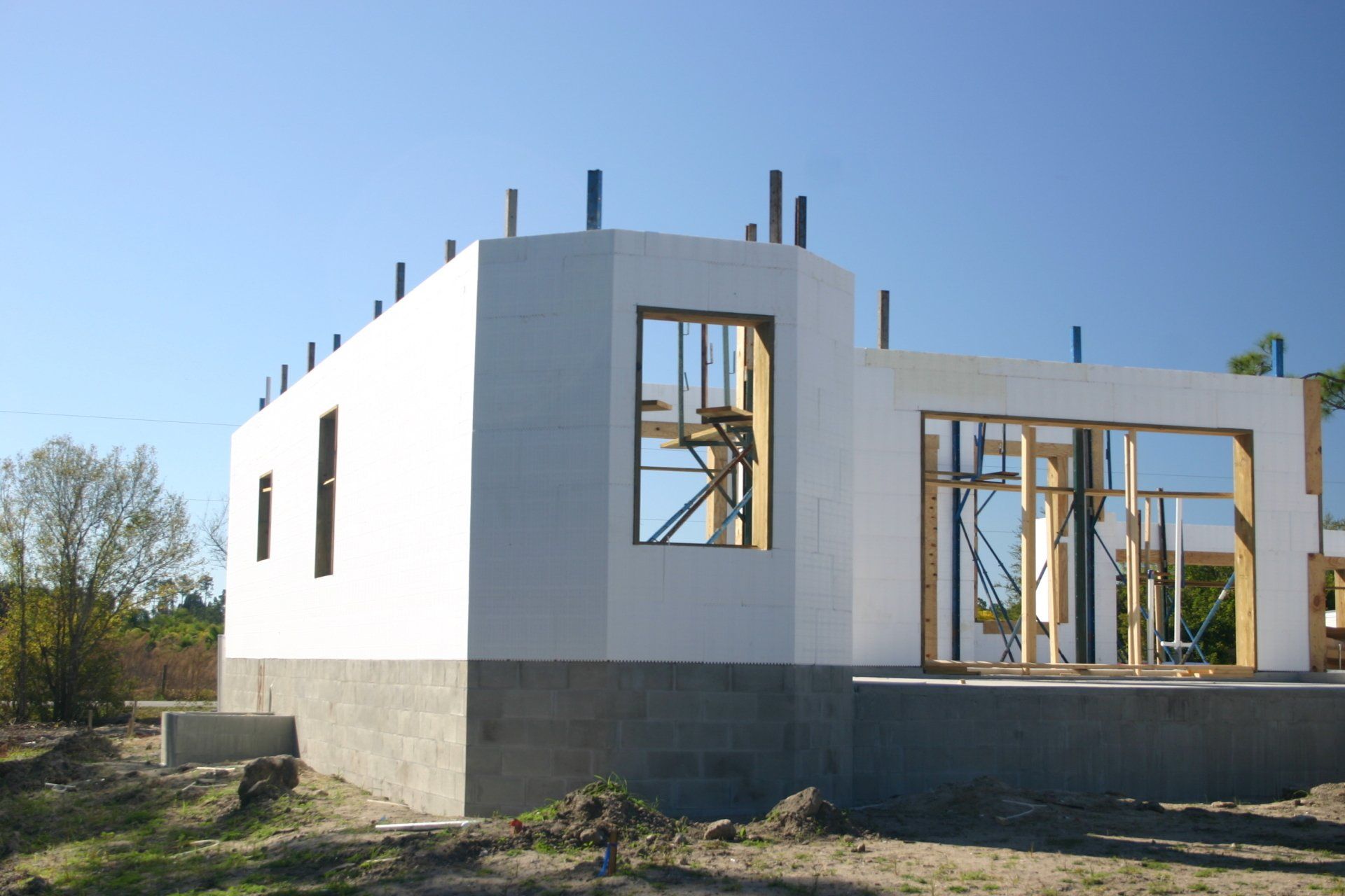 House under construction with white walls, wooden frames, and concrete foundation against a blue sky.