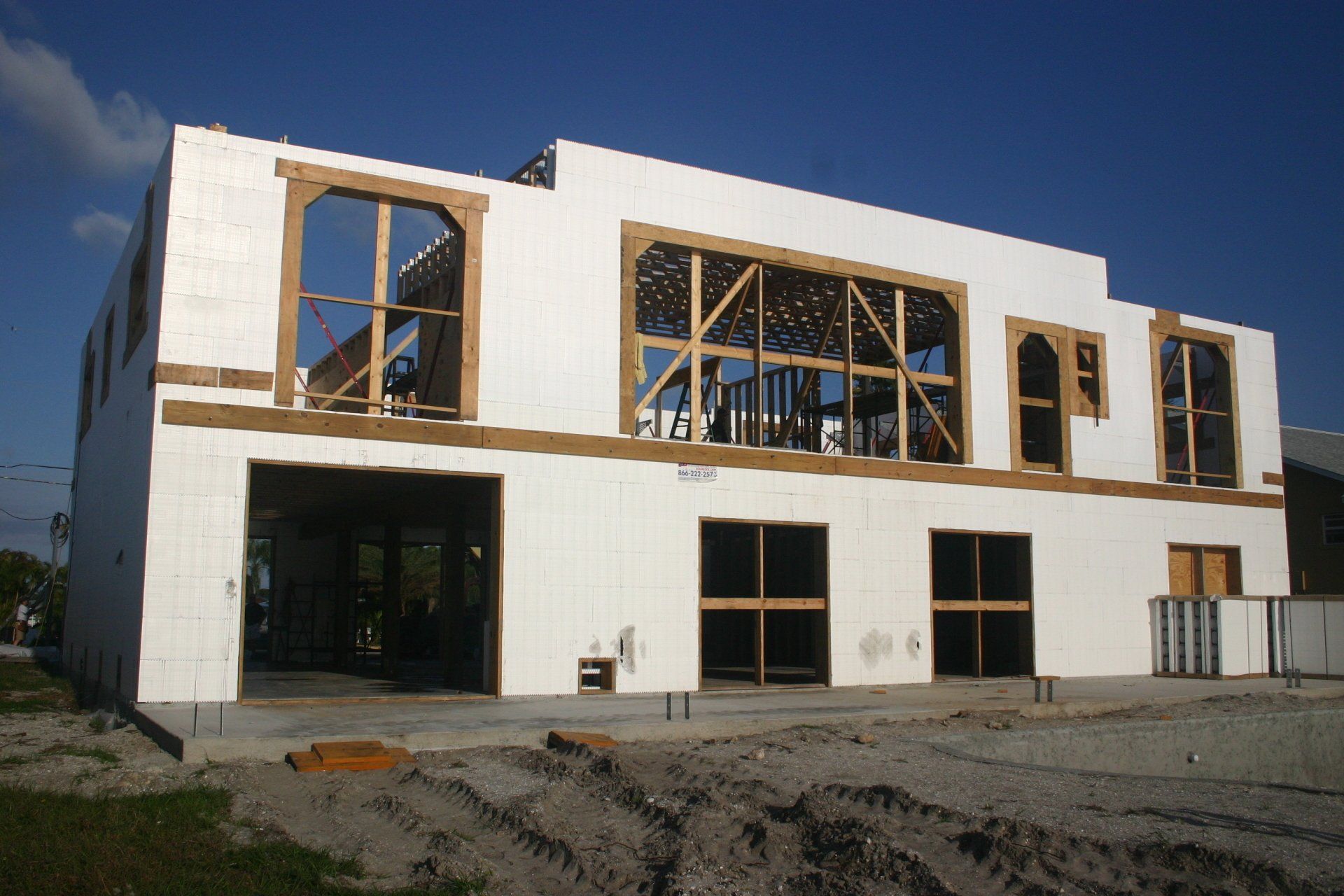 Two-story white building under construction with wooden window frames and open garage door, bright sunny day.