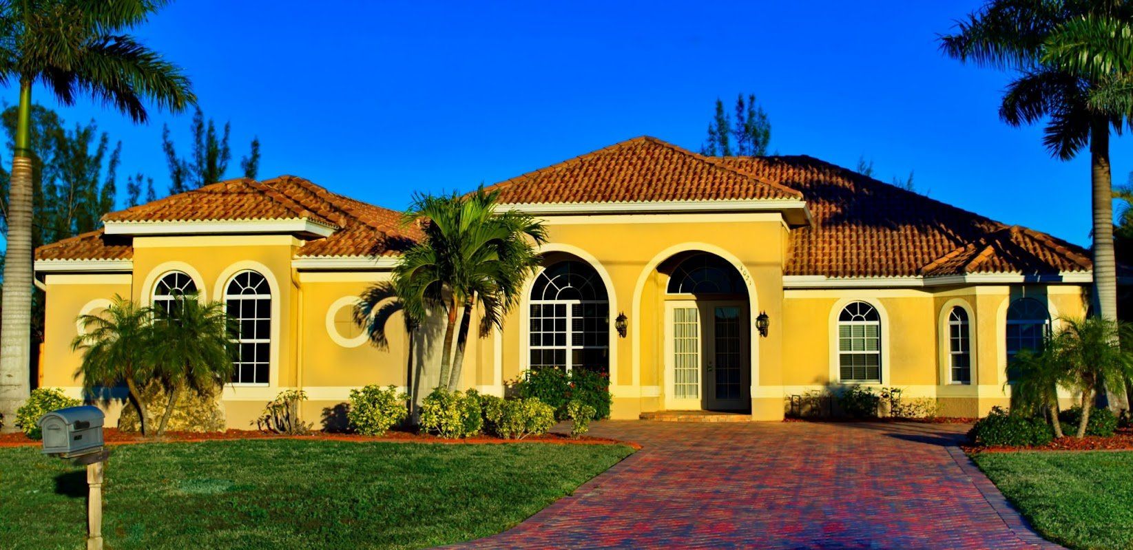 Yellow stucco house with red tile roof and brick driveway. Palm trees, blue sky, and green grass.