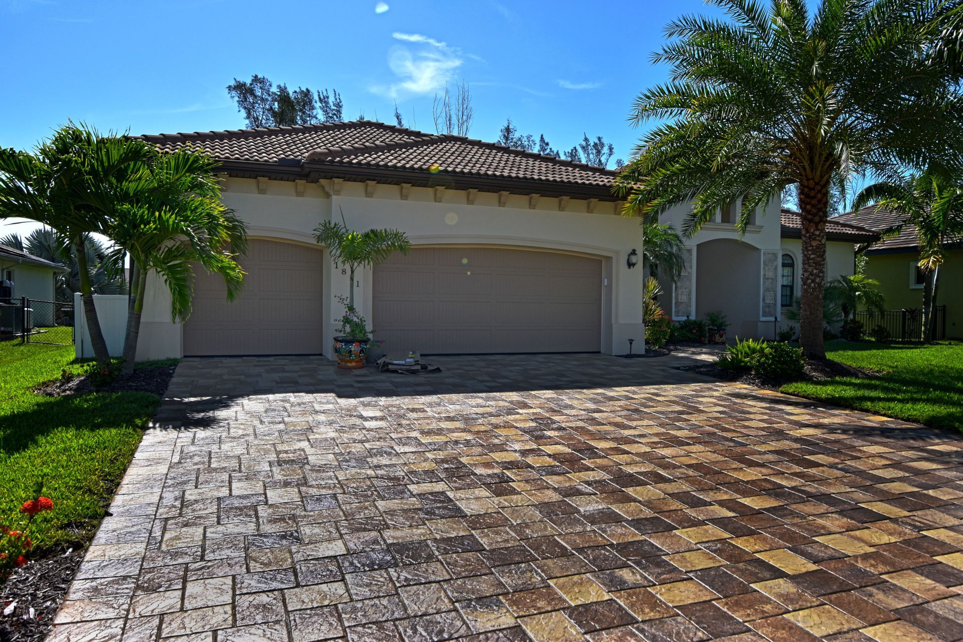 Tan house with a brick driveway and two-car garage under a blue sky. Palm trees in the yard.