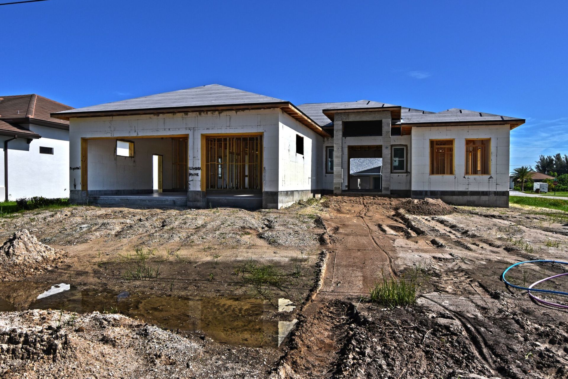 House under construction, exterior view; unfinished concrete walls, exposed framing, dirt yard, bright blue sky.