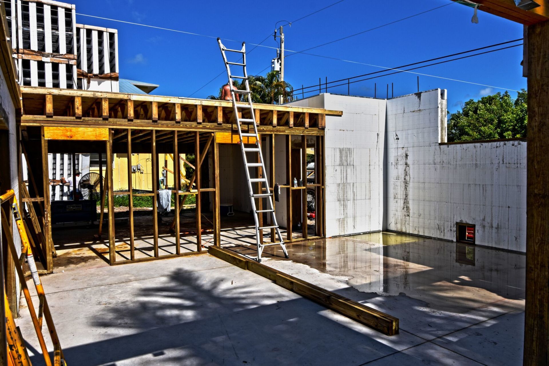 Construction site with wooden frames, ladder, and partially built walls against a bright blue sky.