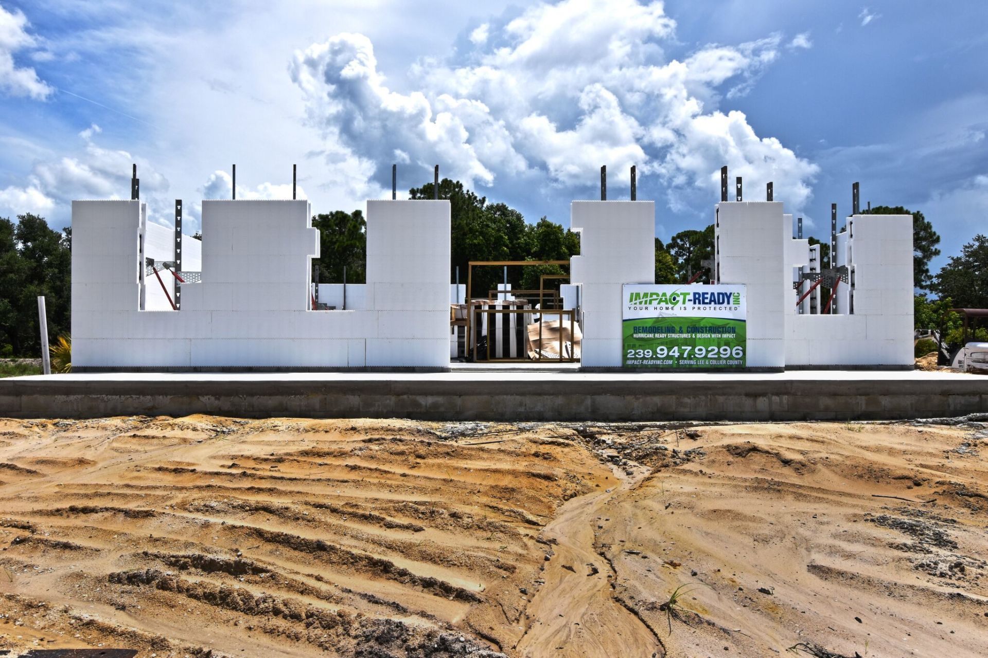 Construction of a white building's framework on a concrete base against a cloudy sky; dirt in foreground.