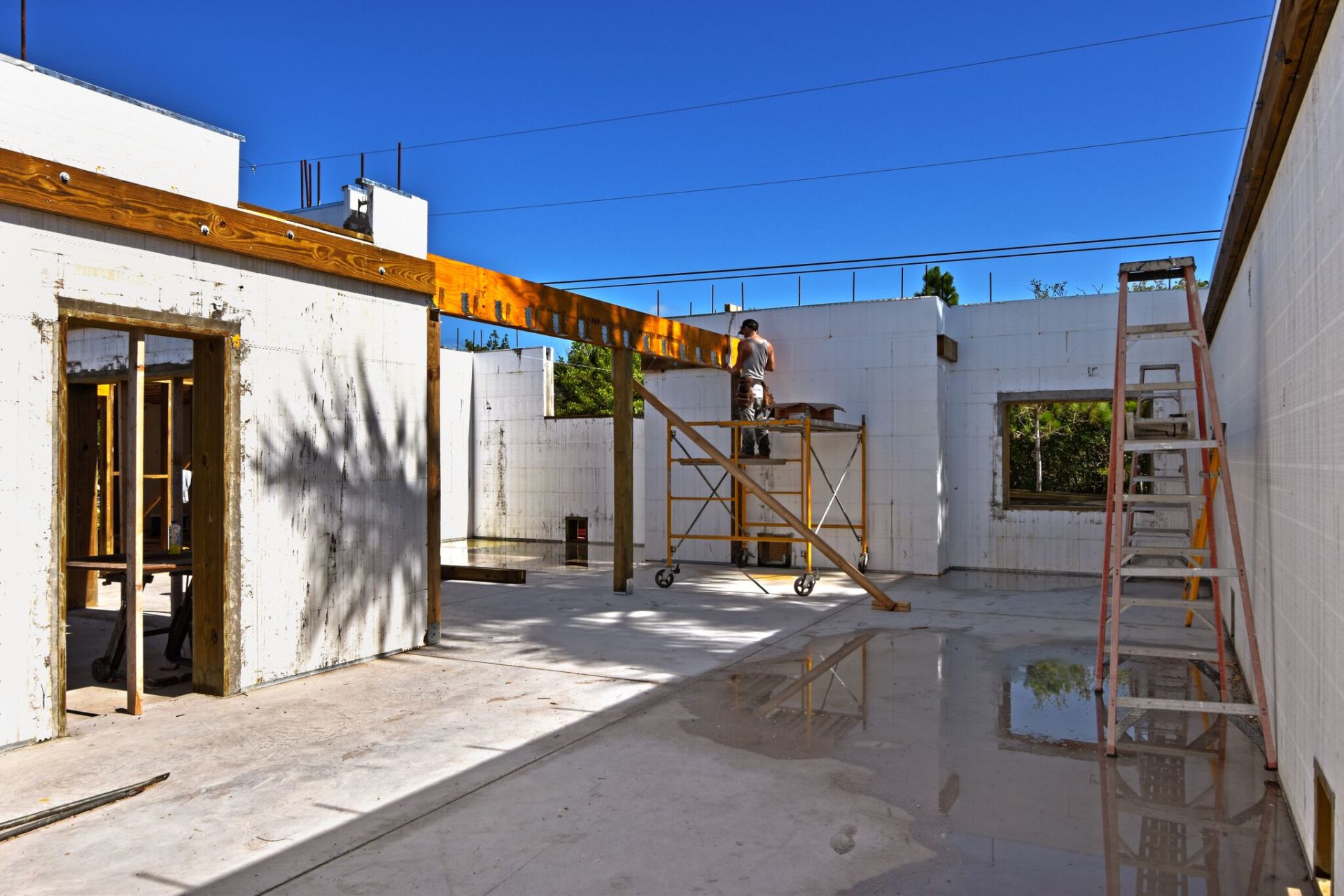 Construction site with white insulated concrete form walls, a worker on scaffolding, ladder, and blue sky.