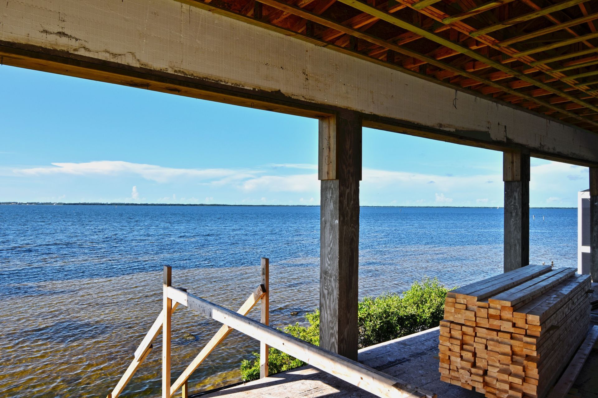 Open-air building under construction overlooking a calm, blue sea. Lumber stacked inside. Bright, sunny day.