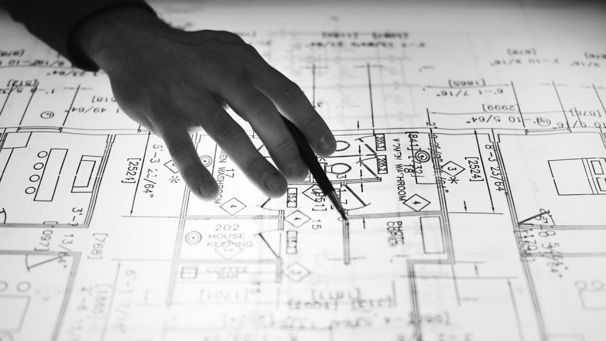 Hand with pen points at architectural blueprints on a light table.