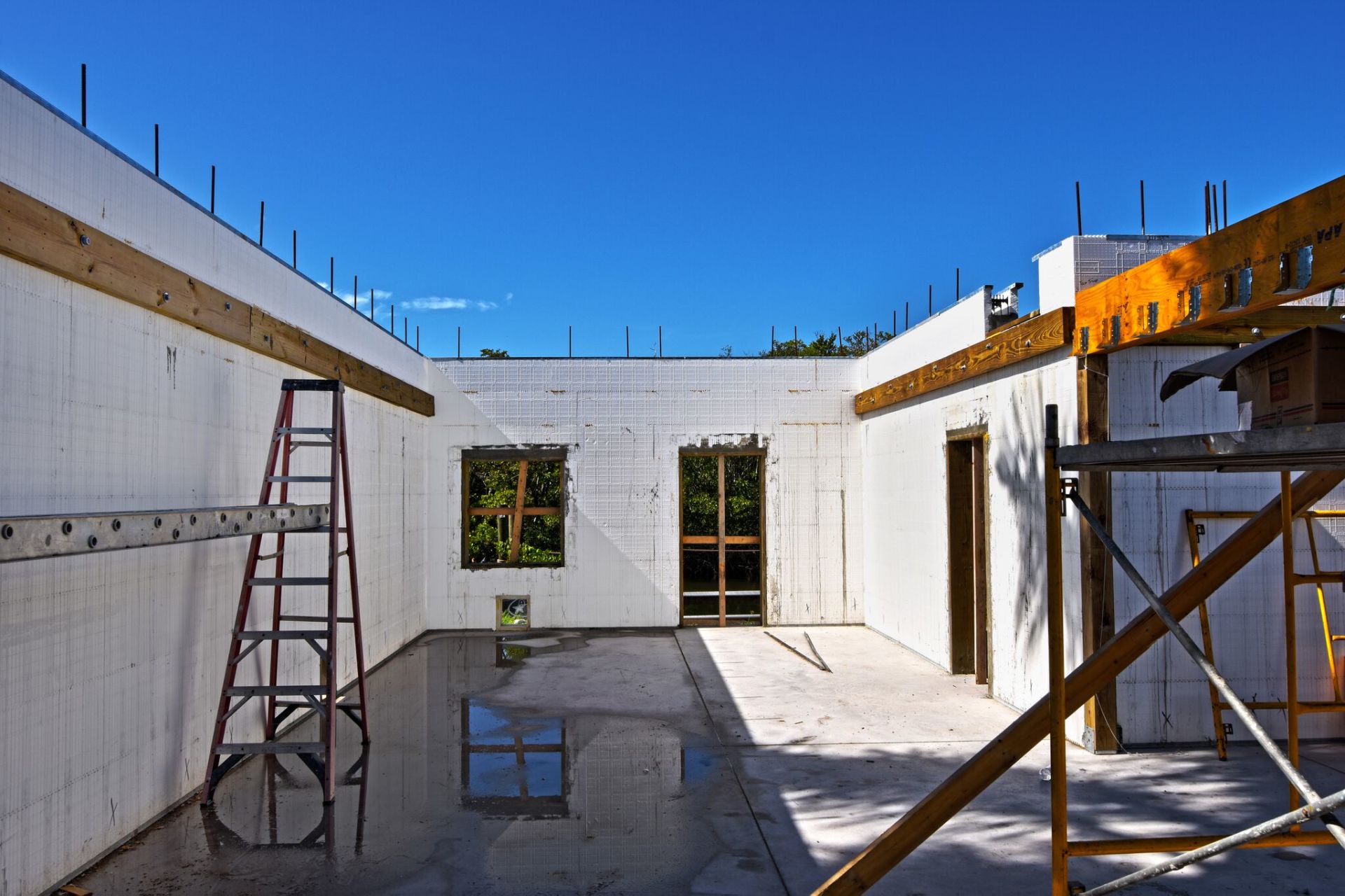 Construction site interior with white insulated concrete forms and a wet floor, a red ladder, and windows.