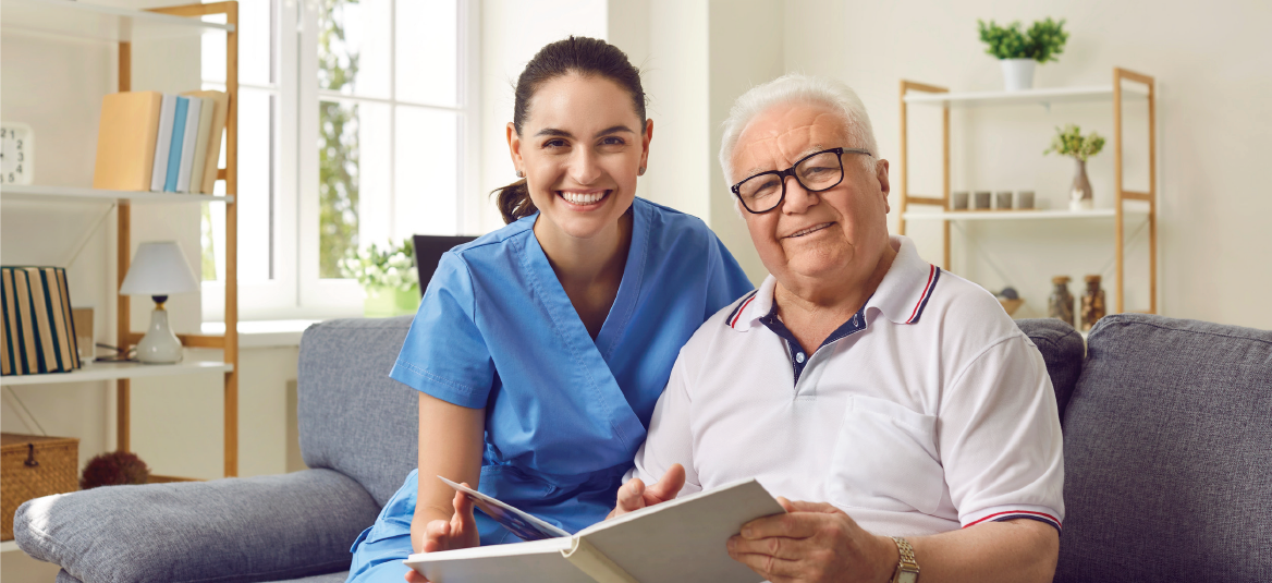 A young woman in blue scrubs smiles next to an older man reading a book on a sofa.