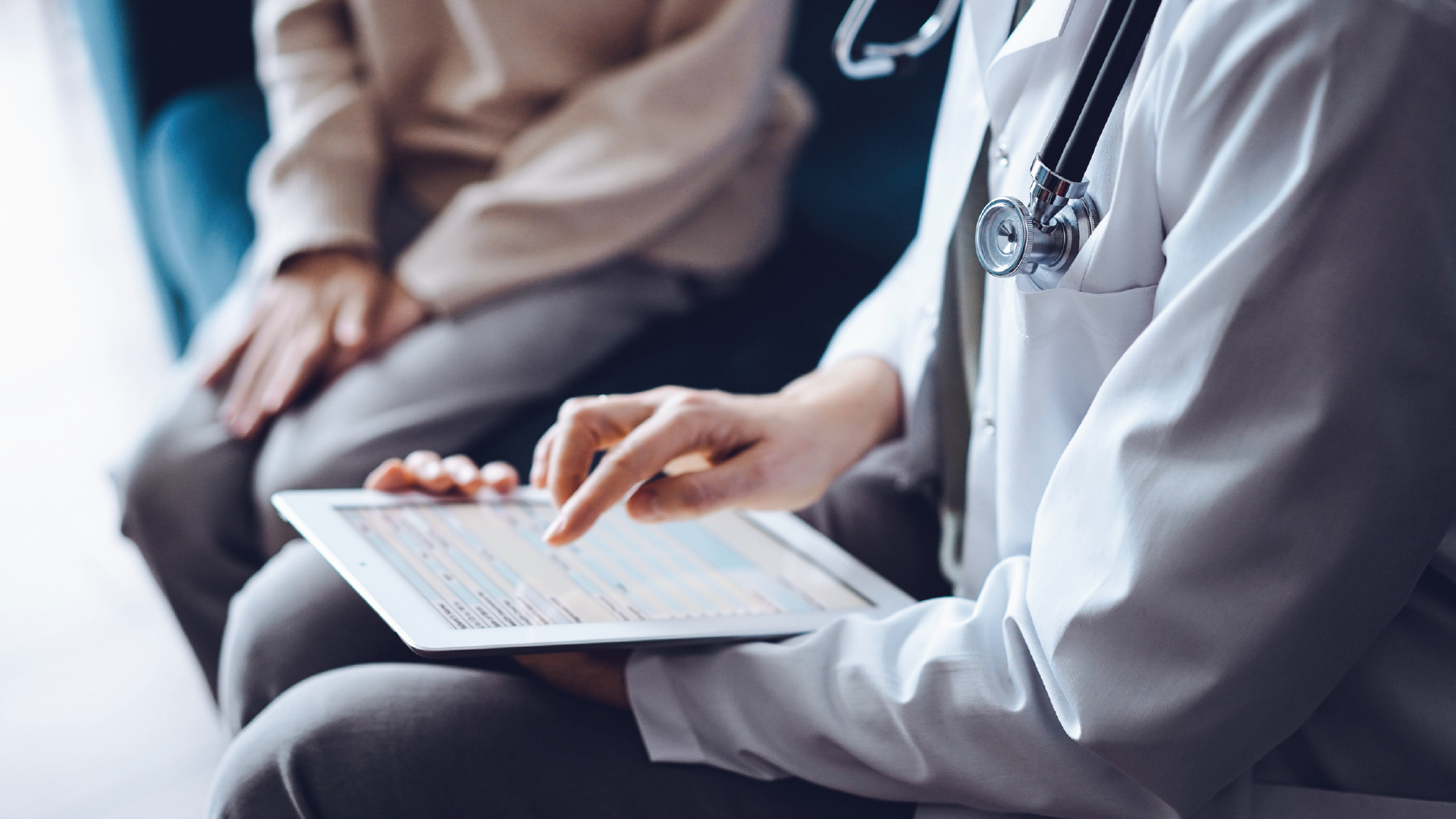 Doctor points at tablet screen, seated with patient. The doctor wears a white coat and stethoscope.