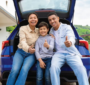Family sitting in the open trunk of a blue car, smiling and giving thumbs up.