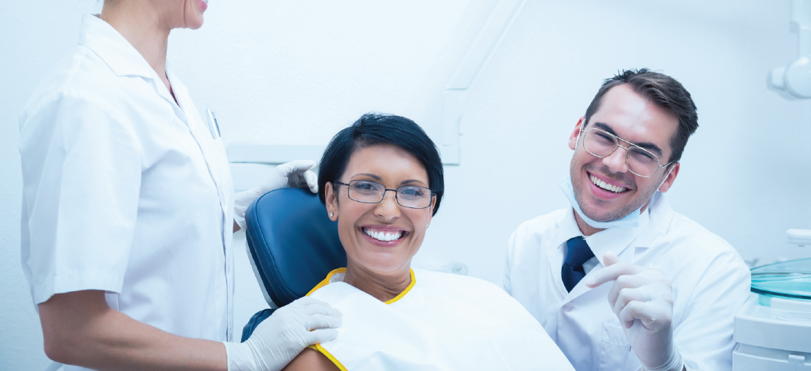 A woman smiling in a dentist's chair. A dentist and assistant smile beside her in a dental office.