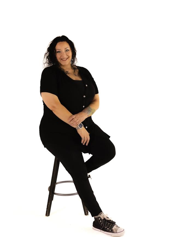 Woman in black outfit sitting on stool, smiling.