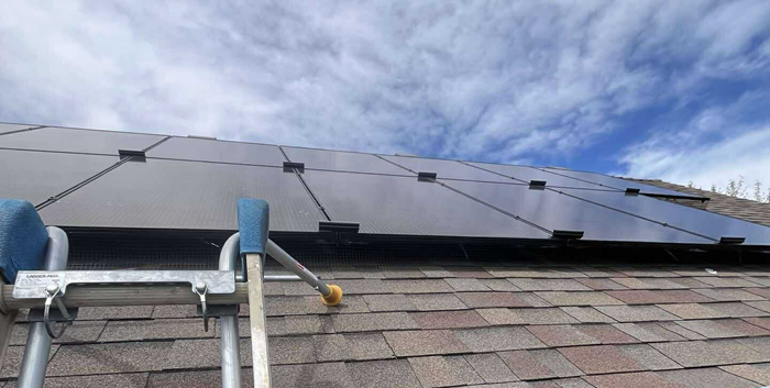 Solar panels installed on a brown shingle roof with a ladder against the side, blue sky in the background.