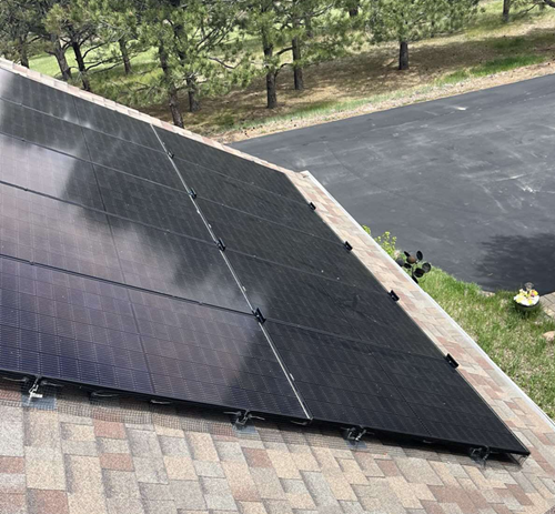 Solar panels on a brown shingled roof, trees and a road in the background.