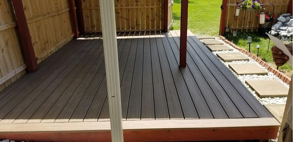 Wooden deck with a brown frame, surrounded by a fence and stepping stones on a sunny day.
