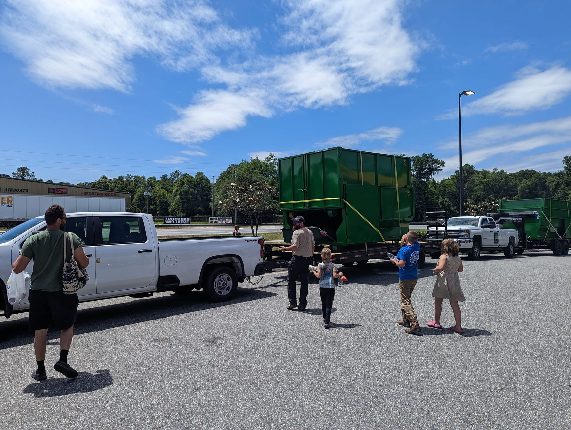 People loading boxes onto green dumpsters on trailers, parked in a gravel lot under a blue sky.