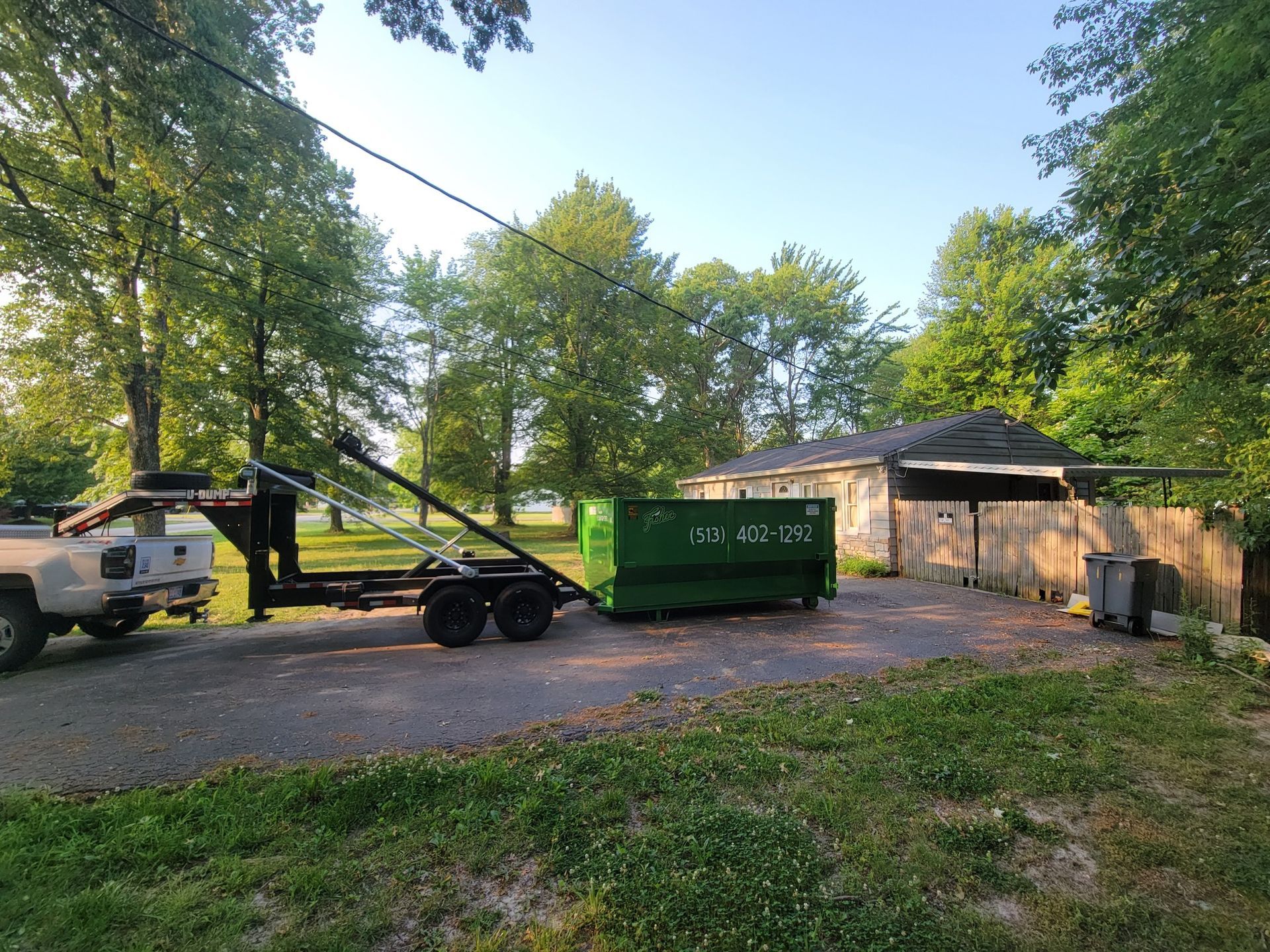 A green dumpster on a trailer parked in front of a small house, trees in the background.