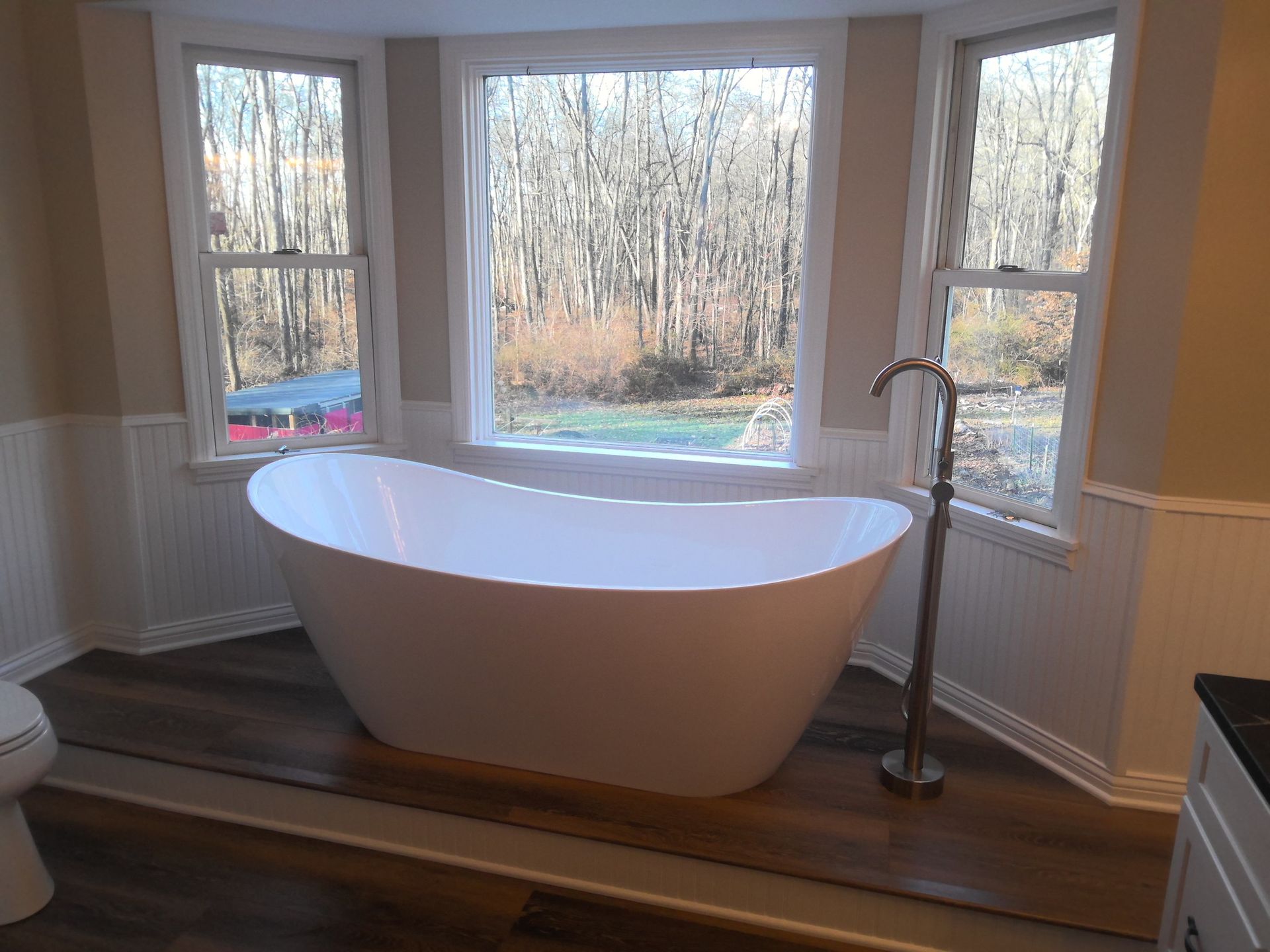 White freestanding bathtub in front of a large window overlooking a wooded area.