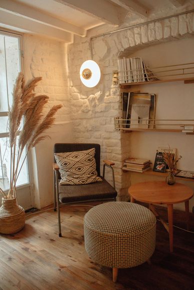 Cozy reading nook: chair, ottoman, small table. Dried plants, white brick walls, and warm lighting create a peaceful space.