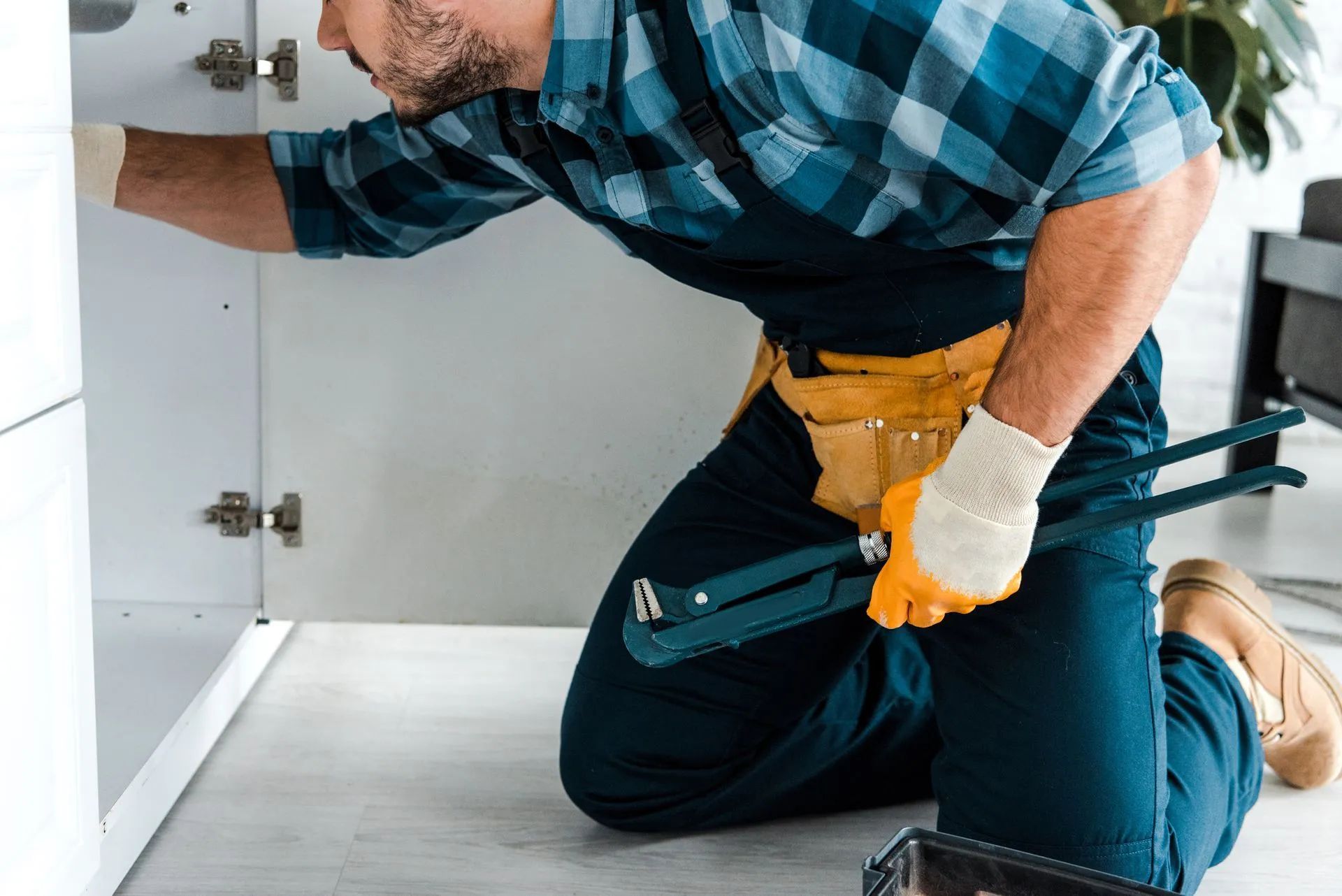 Plumber kneeling under sink, holding a wrench, wearing overalls and gloves.