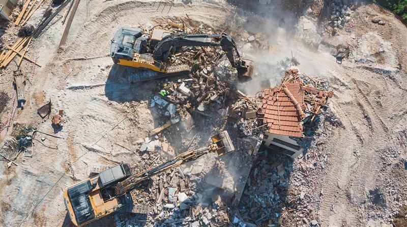 Aerial view of excavators demolishing a building, kicking up dust. Debris scattered across the site.