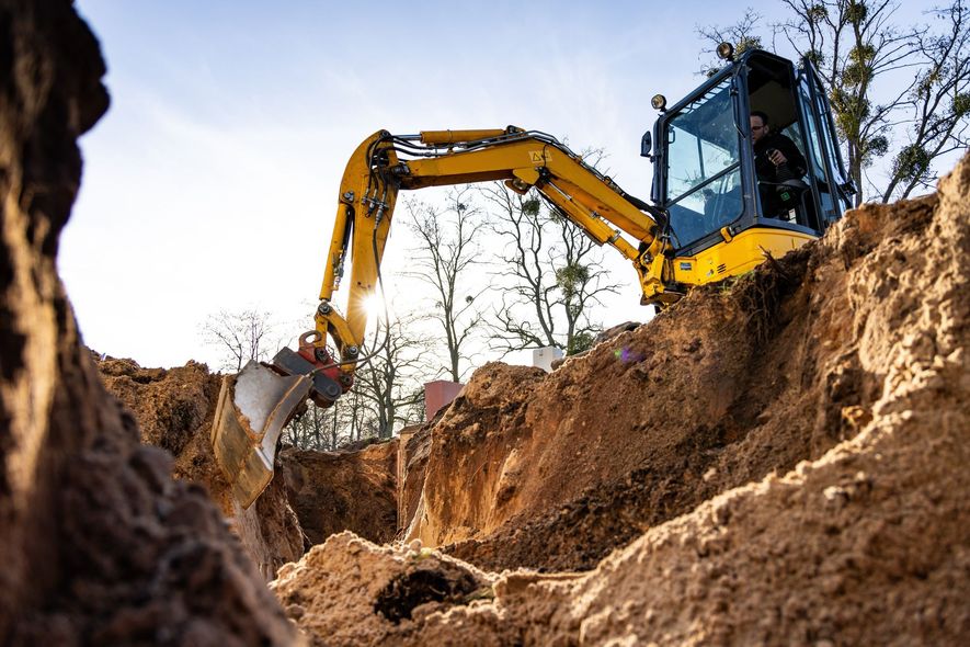 Yellow excavator digging trench in the dirt, with sunlight and trees in the background.