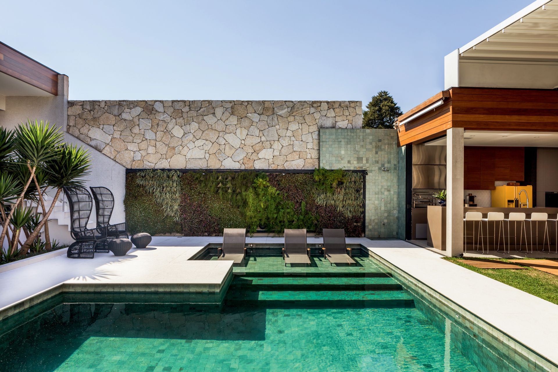 Piscina revestida com hijau, espreguiçadeiras, muro de pedra, jardim vertical e cozinha gourmet.
