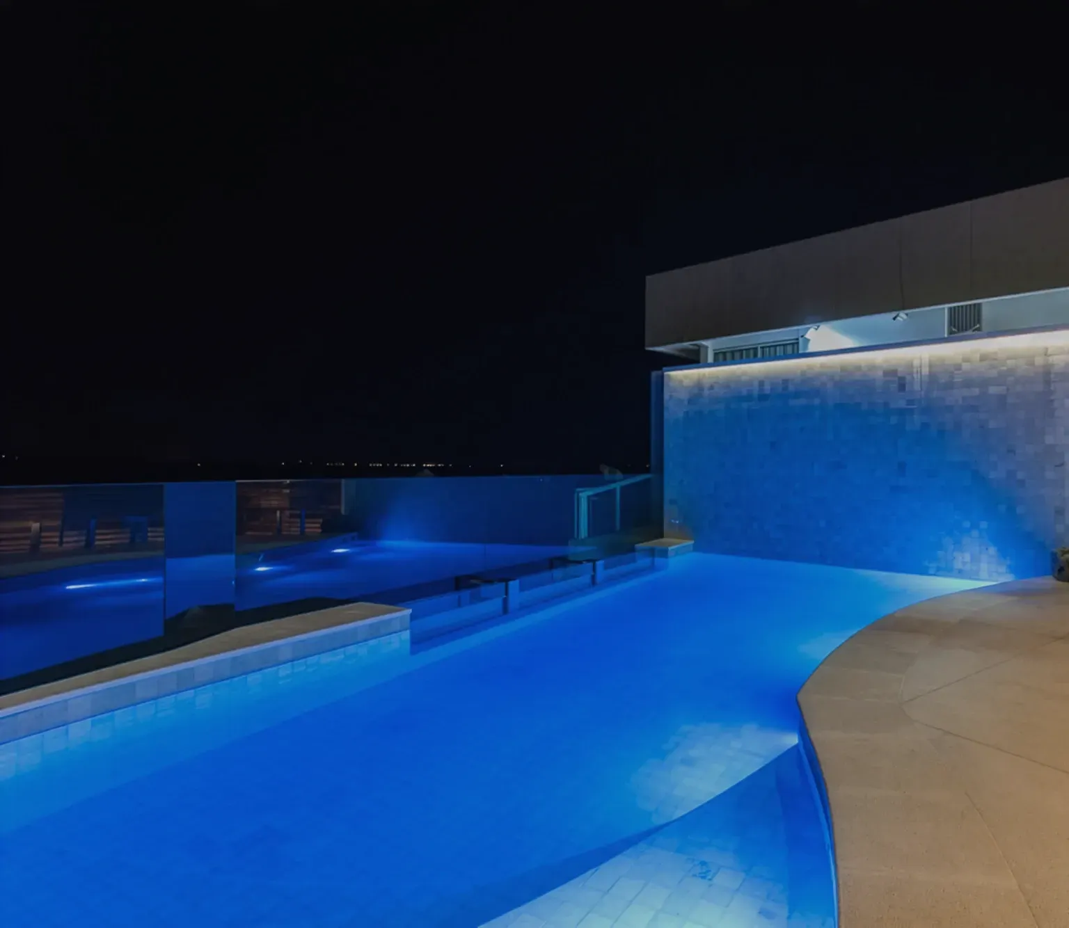 Piscina revestida de pedra água marinha, em cobertura iluminada de azul, à noite.