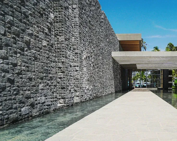 Muro revestido com pedra hitam bruta Artemano ao lado de um espelho de água e um caminho que leva à entrada de um prédio.