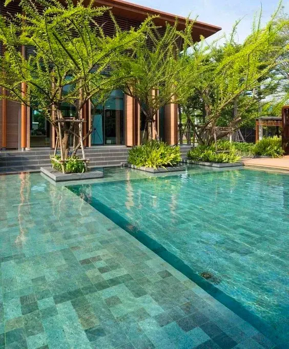 Piscina revestida com hijau Artemano em frente a uma casa de alto padrão com árvores e plantas.