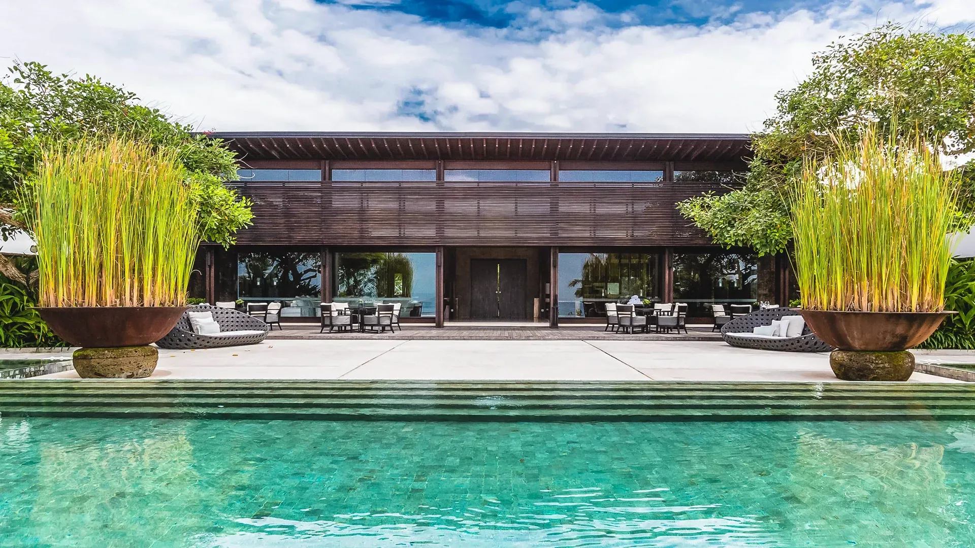 Casa de alto padrão com piscina revestida com Hijau Artemano, paisagismo exuberante e área de estar externa sob um céu azul.