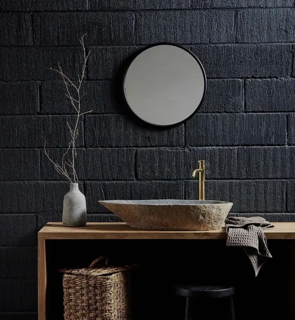 Parede de tijolos escuros com espelho redondo, cuba de pedra natural sobre bancada de madeira, torneira de latão, cesto.