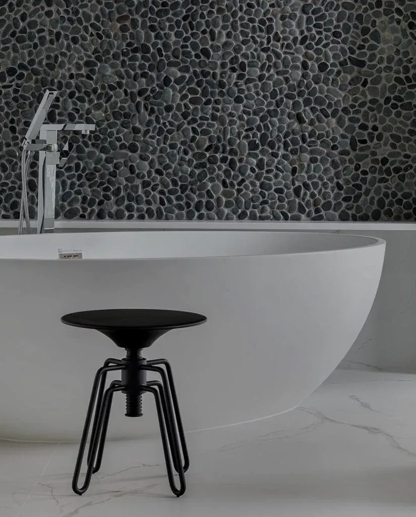 Banheira branca com banquinho preto em frente; parede revestida com seixos pretos.