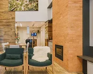 Sala de estar moderna com parede e lareira revestidos com tijolo inglês, duas poltronas verdes e uma área de jantar ao fundo.