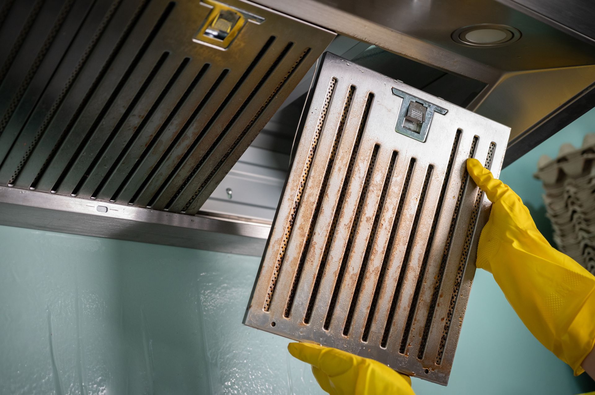 Yellow-gloved hands hold a metal vent filter under a kitchen range hood.