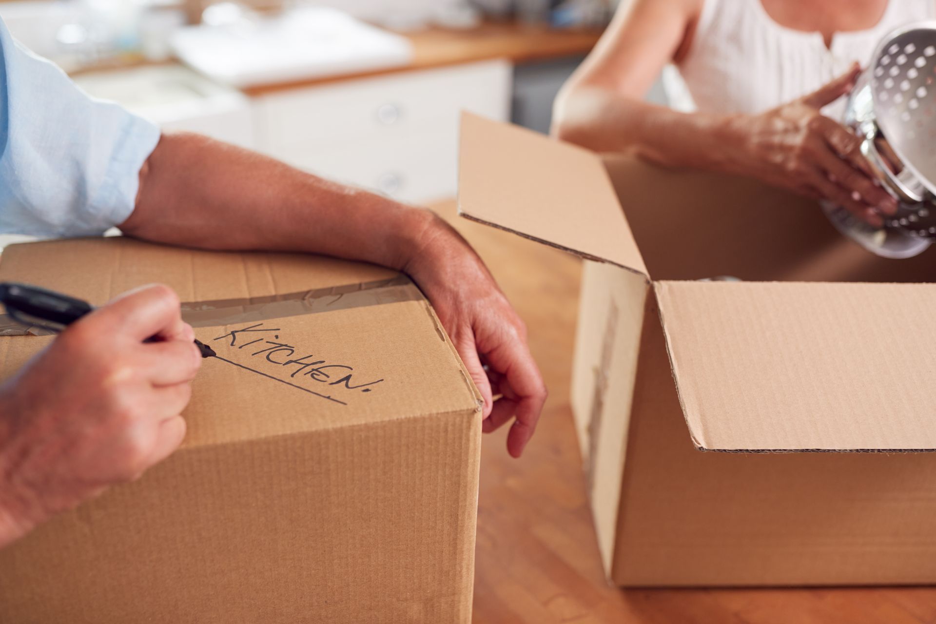 Person writing on a cardboard box while another person packs a kitchen item into another box, possibly moving.