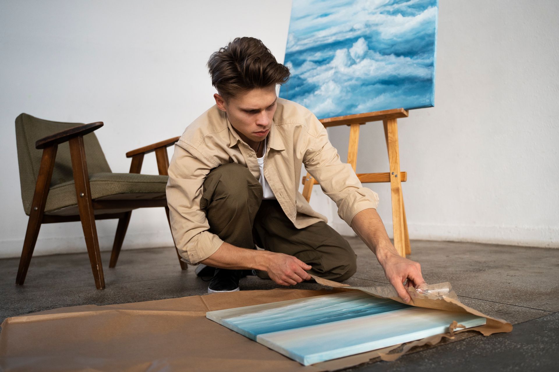 A man is kneeling on the floor looking at a painting.