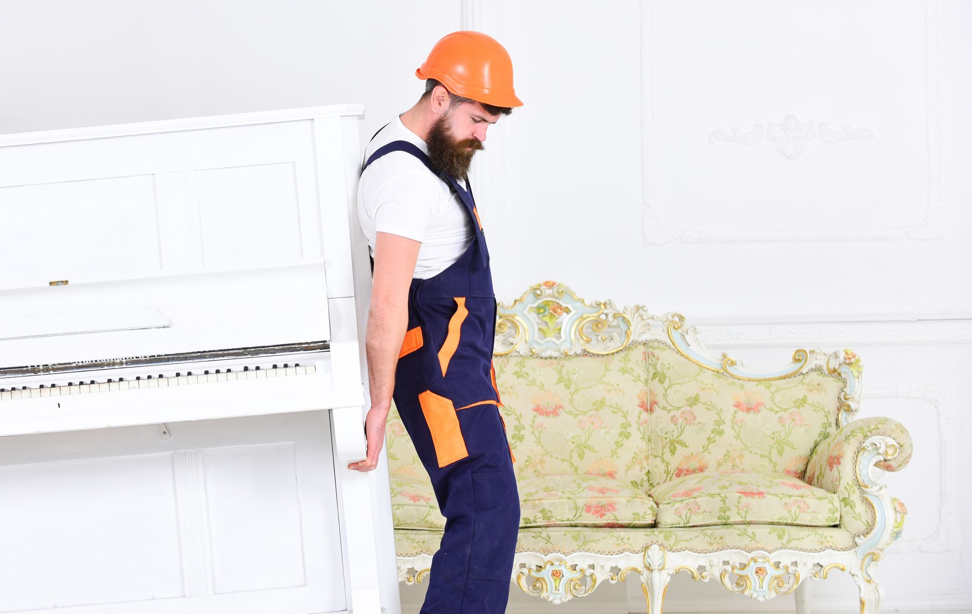 A man in overalls and a hard hat is standing next to a piano in a living room.
