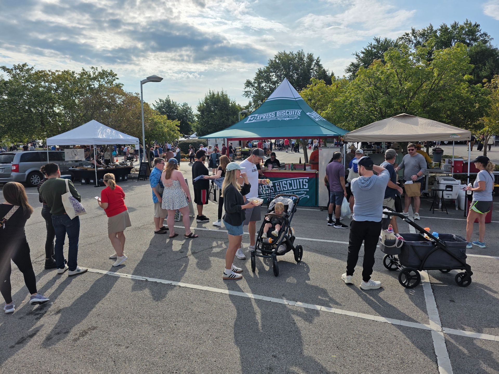 Outdoor food market with people, tents, and food vendors in a parking lot. Cloudy sky and trees in the background.