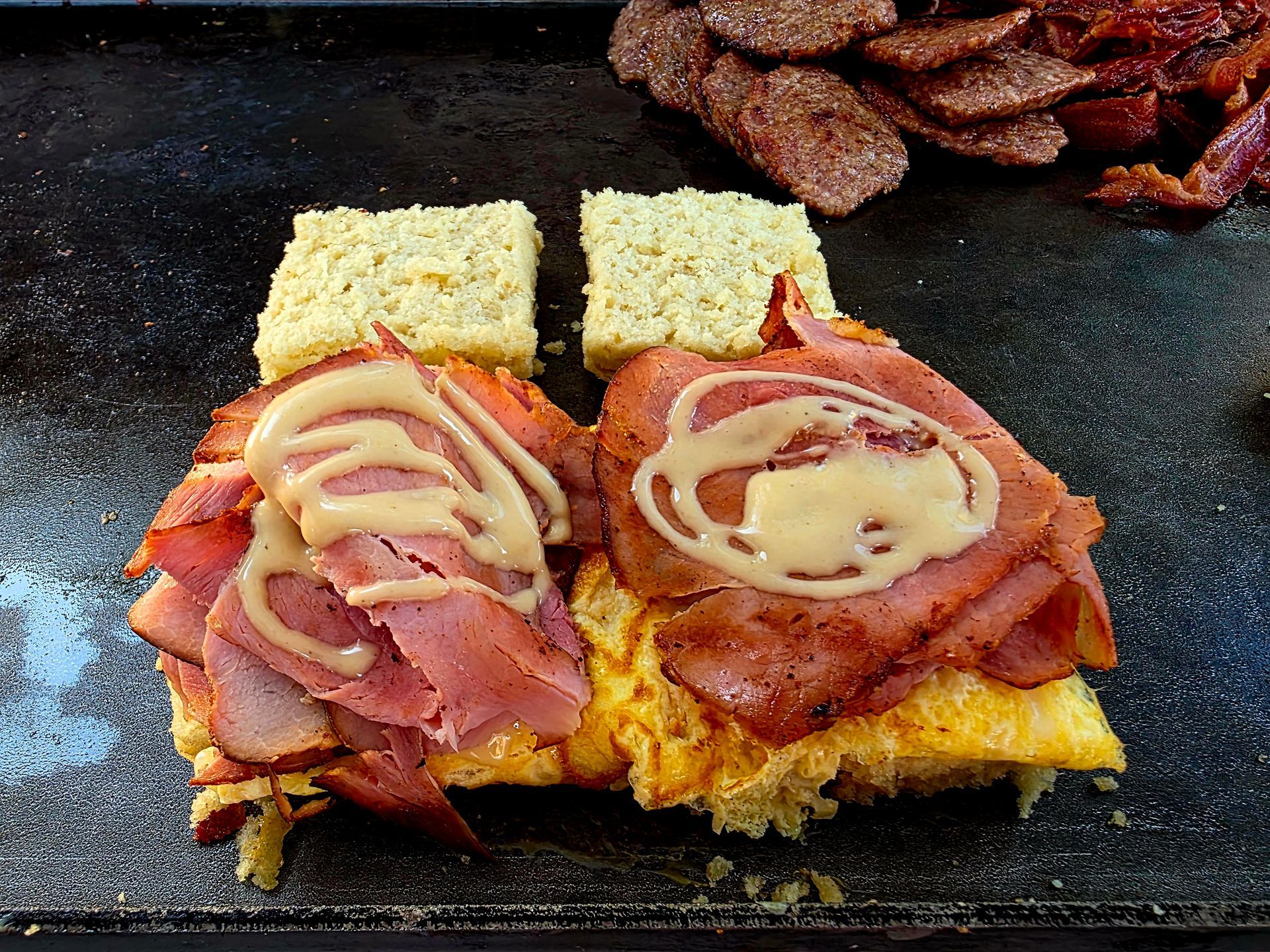 Sandwich being assembled on a griddle, featuring ham, sauce, and bread.