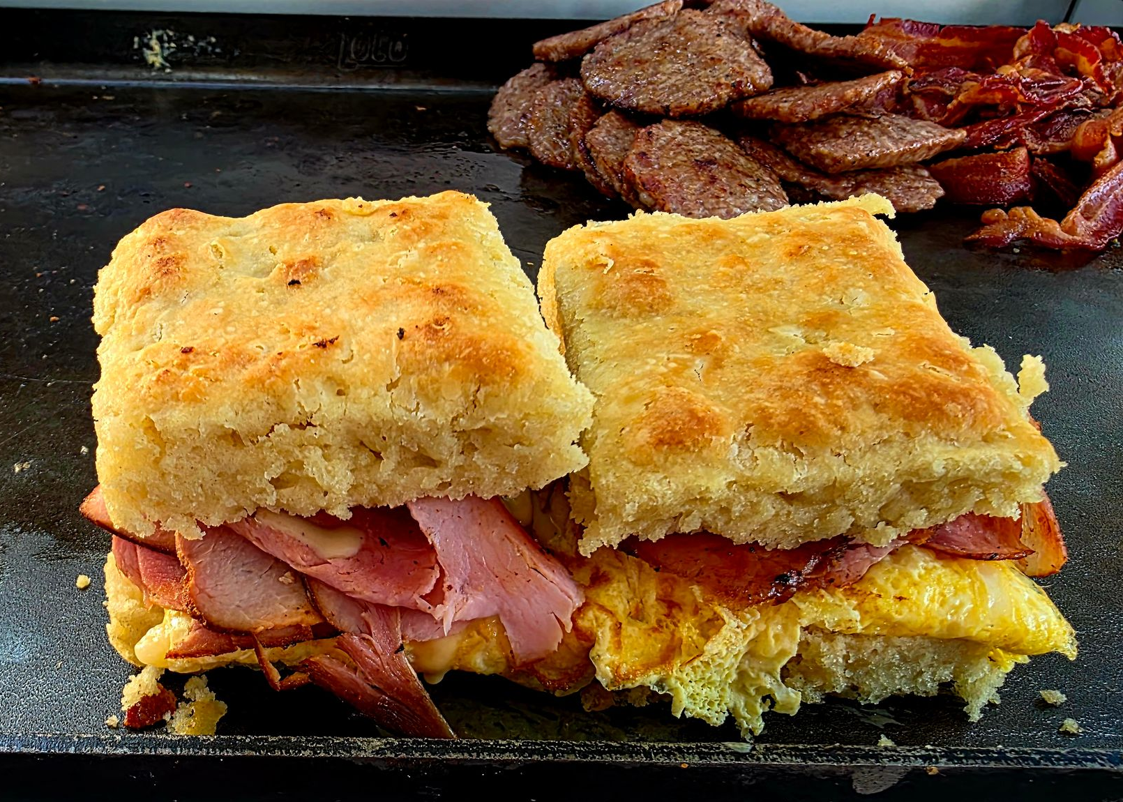 Breakfast sandwich with egg, bacon, and ham on a biscuit; sausage and bacon in background.